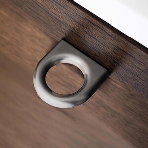 May include: A close-up of a contemporary, brushed metal drawer pull with a circular handle. The pull is mounted on a dark brown wooden surface, with a white edge and a glimpse of a book in the background.