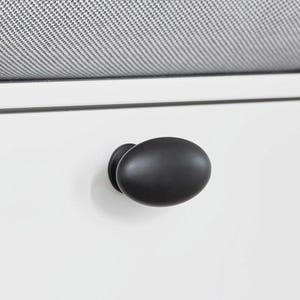 May include: A white cabinet with a black, oval-shaped knob. The knob is centrally positioned on the cabinet door. A grey and white patterned cushion is visible above the cabinet.