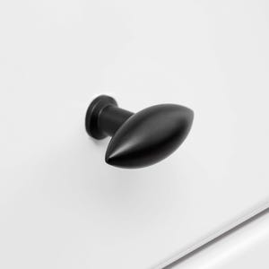 May include: A black cabinet knob with a teardrop shape, mounted on a white cabinet door. The knob has a matte finish and a simple, modern design. The image is a close-up.
