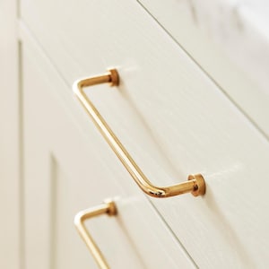 May include: Close-up of two gold-coloured cabinet pulls with a curved, bar-like design. The pulls are mounted on a light-coloured cabinet door. The cabinet door is made of wood and has a simple, recessed panel design. The countertop is white marble.