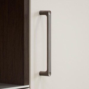 May include: A dark grey cabinet pull handle with a rectangular design. The handle is mounted on a white cabinet door, with a dark brown cabinet frame visible on the left. The handle has rounded edges.