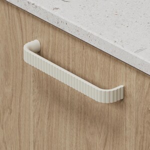 May include: A cream-coloured cabinet pull with a ribbed design is mounted on a light wood cabinet. The pull has a curved shape and is positioned near a white speckled countertop. The cabinet door has a natural wood grain.