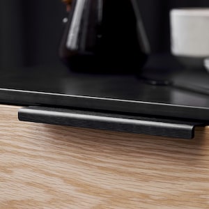 May include: A black drawer pull with a woodgrain finish. The pull is mounted on a light wood drawer front.