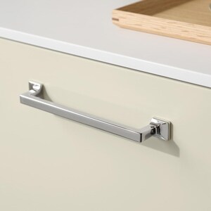 May include: A polished chrome drawer pull with a rectangular bar and square end mounts. The hardware is mounted on a cream-coloured cabinet door. A wooden tray and a cup are visible in the background.