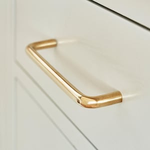 May include: Close-up of a gold-coloured cabinet pull handle. The handle is rectangular with rounded corners and is mounted on a white cabinet door. The hardware has a shiny, metallic finish.