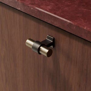 May include: A close-up of a cabinet door with a decorative handle. The handle is a combination of black and gold, with a cylindrical design. The cabinet door is a dark brown colour, and the countertop is a reddish-brown marble.