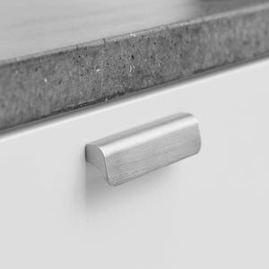 May include: A close-up of a brushed metal cabinet pull handle. The handle is mounted on a white cabinet door, with a grey countertop in the background. The handle has a sleek, minimalist design.