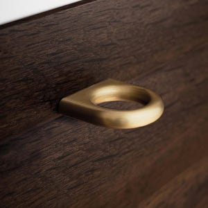 May include: A close-up of a brass-coloured ring pull handle on a dark brown wooden surface. The ring pull has a simple, modern design, and the wood grain is visible.