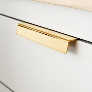 May include: A close-up shot of a white drawer featuring a gold-coloured handle. The handle is rectangular and has a modern design. The drawer is part of a larger piece of furniture, with a light-coloured wooden top visible.