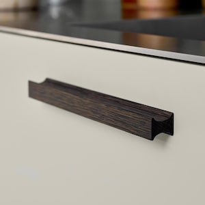 May include: A dark brown wooden cabinet pull with a grooved design. The rectangular handle is mounted on a cream-coloured cabinet door. The kitchen hardware is made of wood and has a modern aesthetic.