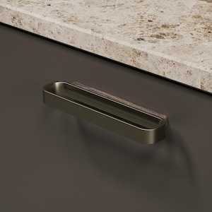 May include: A bronze-coloured cabinet pull with a rectangular design. The pull is mounted on a dark brown cabinet door, with a light-coloured countertop in the background. The pull has a sleek, modern aesthetic.