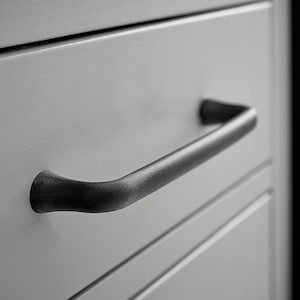 May include: A black metal drawer pull with a curved design. The pull is mounted on a white drawer front.