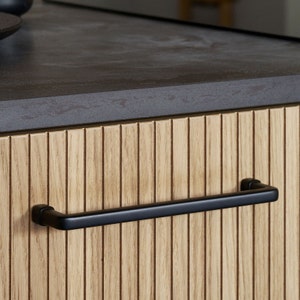 May include: Black cabinet door handle with a rectangular shape and rounded edges. The handle is mounted on a wooden cabinet door with vertical grooves.