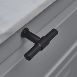May include: A close-up shot of a black T-bar cabinet pull. The pull is attached to a grey cabinet door, with a white marble countertop in the background. The hardware has a matte finish.