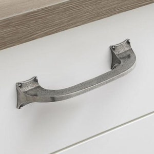May include: A silver-toned drawer pull with a curved design and pointed ends. The pull is mounted on a white cabinet door.