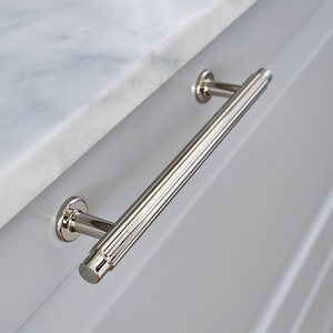 May include: A polished nickel cabinet pull with a fluted design. The pull is mounted on a white cabinet door.
