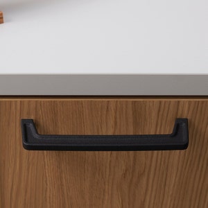 May include: Black cabinet pull with a slightly curved rectangular shape. The pull is mounted on a wooden cabinet door.