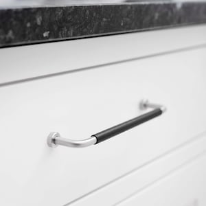 May include: Close-up of a white cabinet drawer with a silver handle. The handle has a curved design with a black grip in the centre. The cabinet is below a dark countertop.