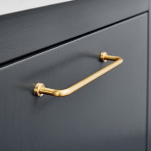 May include: A gold-coloured cabinet pull handle with a curved bar design. The handle is mounted on a dark grey cabinet door. The hardware has a cylindrical base and a smooth, modern aesthetic.