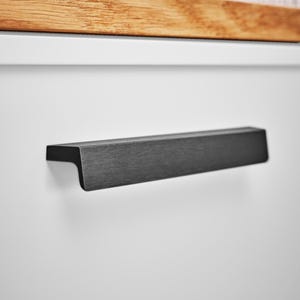 May include: A close-up of a black metal cabinet pull handle. The handle is rectangular with a slightly curved edge and is mounted on a white cabinet door. A wooden surface is visible above the handle.