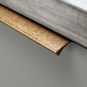 May include: Close-up of a section of a cabinet or drawer. The image shows a bronze-coloured metal pull handle with a curved end, positioned below a grey, textured surface. The handle has a distressed finish.