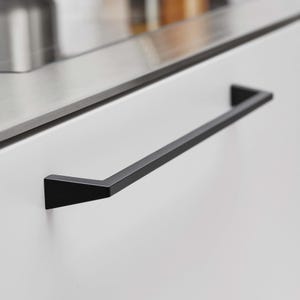 May include: A black, rectangular cabinet pull with a minimalist design. The pull is mounted on a white cabinet door, with a stainless steel countertop visible in the background. The hardware has a modern, sleek aesthetic.