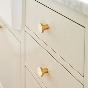 May include: Close-up of a light-coloured cabinet with three drawers. Each drawer has a round, gold-coloured knob. The cabinet has a simple, modern design, with visible wood grain.