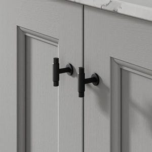 May include: Close-up of a light grey cabinet with two black T-shaped handles. The cabinet doors have a recessed panel design. A white countertop with grey veining is visible above the cabinet.