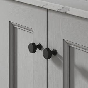 May include: A light grey cabinet with two black, round knobs. The cabinet doors have a recessed panel design. A white countertop with grey veining sits above the cabinet.