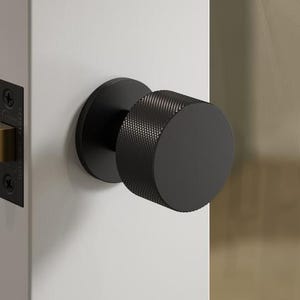 May include: A black door knob with a textured cylindrical design. The knob is mounted on a white door, with a black door plate and a gold-coloured latch visible. The door is partially open, revealing a glimpse of the interior.