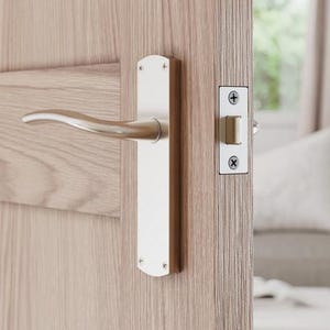 May include: A light oak wooden door with a silver-coloured handle and matching door latch. The door is ajar, offering a view of a living room with a sofa and white cushions.