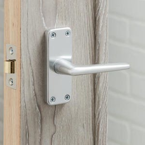 May include: A close-up of a door featuring a silver handle and a light wood grain finish. The handle has a rectangular backplate and a curved lever. The door frame is light wood. The door is ajar, showing a white brick wall.