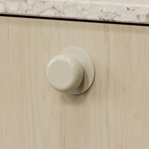 May include: A light beige cabinet knob with a circular base and a rounded, cylindrical handle. The knob is mounted on a light-coloured wooden cabinet door. The countertop above is a speckled beige and grey.