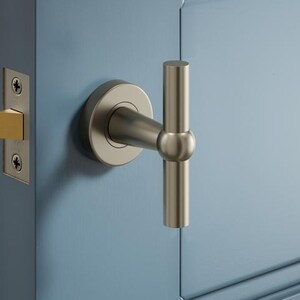 May include: A close-up of a door handle and latch. The handle has a brushed nickel finish with a T-bar design. The door is painted a light blue colour. The latch plate is also brushed nickel with a gold-coloured latch.