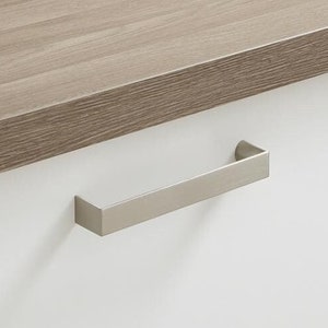 May include: A brushed nickel cabinet pull with a rectangular shape. The pull is mounted on a white cabinet door.