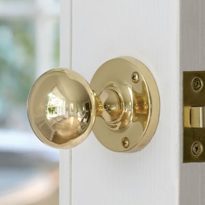 May include: A close-up of a polished brass door knob and latch set. The round knob reflects the surrounding environment. The set is mounted on a white door, with a glimpse of a window in the background.