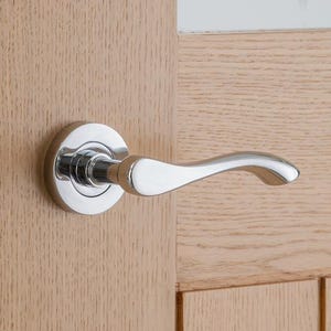 May include: A shiny chrome door handle with a curved lever is fixed to a light-coloured wooden door. The door also has a matching chrome door latch. The door is slightly ajar, showing a glimpse of a white wall.