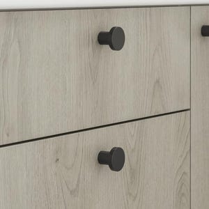 May include: Close-up of a light grey wooden cabinet with black round knobs. The cabinet has three drawers and a door. The wood grain is visible on the surface. The knobs are evenly spaced.