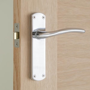 May include: A shiny chrome door handle and plate are fixed to a light-coloured wooden door. The handle has a curved design, and the plate is rectangular with rounded corners and decorative screws.