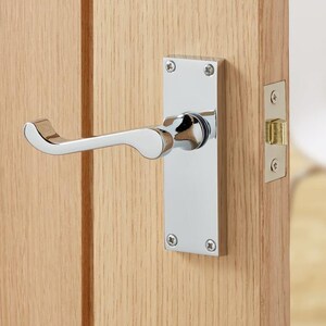 May include: A polished chrome door handle and rectangular backplate are fixed to a light-coloured wooden door. The handle features a curved lever design. The door is ajar, showing a room with white chairs.