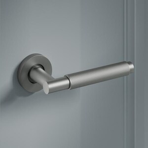 May include: A brushed nickel door handle with a textured grip and a round backplate. The handle is mounted on a grey door, with the latch plate visible. The design is modern and minimalist.