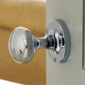 May include: A shiny chrome door knob, featuring a rounded, oval handle and a circular backplate. The door knob is fixed to a light-coloured door, with a glimpse of a beige sofa in the background. The hardware reflects light.