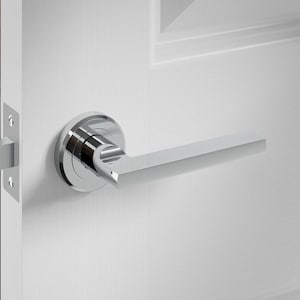 May include: A polished chrome door handle and latch set. The handle has a sleek, modern design with a rectangular lever. The door is white with recessed panels. The latch plate is also chrome.