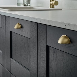 May include: Close-up of a kitchen island with dark grey cabinets and gold-coloured hardware. The countertop is light grey with a sink and tap visible in the background. The cabinet doors have a wood grain texture.