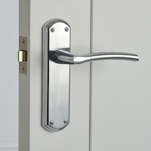 May include: A polished chrome door handle with a curved lever and a rectangular backplate. The handle is mounted on a white door, with visible screws securing the backplate. The door frame and latch are also visible.