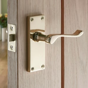 May include: A polished brass door handle and latch set fixed to a light-coloured wooden door. The handle features a curved lever design, and the rectangular backplate is secured with four screws. The door is partially open, showing a glimpse of the interior.