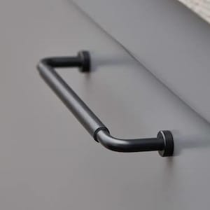 May include: A black metal cabinet pull handle with a curved design. The handle is mounted on a grey cabinet door. The handle has a cylindrical bar and two circular mounting points.