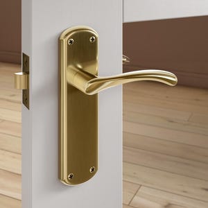 May include: A brass-coloured door handle and backplate are fixed to a white door. The handle has a curved design, and the backplate is rectangular with rounded edges. Four screws secure the plate. The door is ajar, showing a wooden floor.