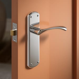 May include: A polished chrome door handle and plate are mounted on a peach-coloured door. The handle has a curved lever design. The door latch is visible on the left side of the door. The door is closed.