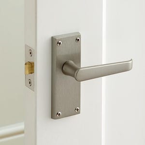 May include: A brushed nickel door handle and plate are mounted on a white door. The rectangular plate has rounded corners and is secured with four screws. The lever handle extends horizontally. A door latch with a gold-coloured strike plate is visible.